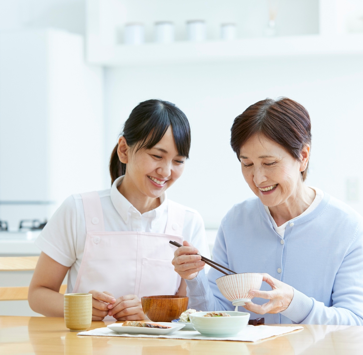 Meal Preparation An Asian senior woman and an Asian caregiver sit together at a dining table, smiling as they enjoy a homemade meal. The atmosphere is warm and friendly, reflecting a moment of shared connection and meal preparation in Gulf Shores.