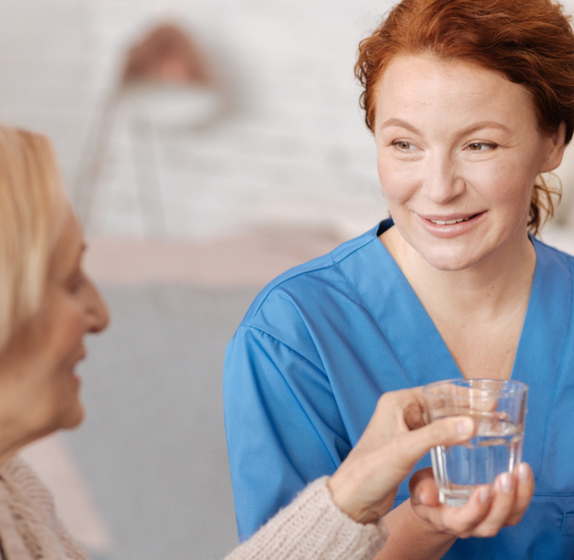 A female caregiver in blue scrubs provides medication supervision in Gulf Shores, handing water and medicine to a senior woman in a warm home setting.