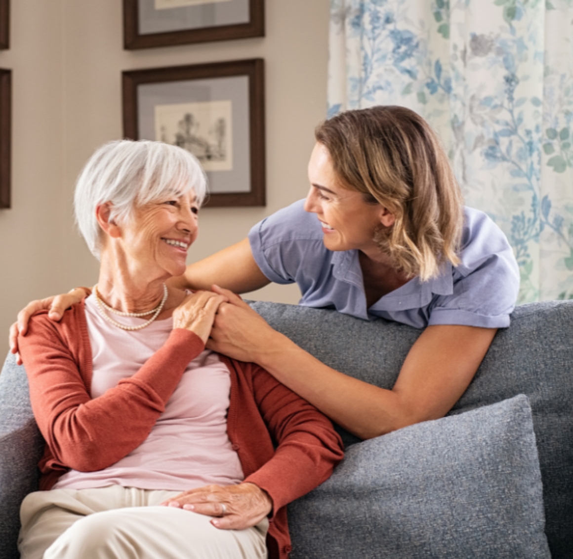 personal care (3) A female caregiver gently puts her arm around a smiling senior woman seated on a sofa, creating a warm and supportive moment that reflects compassionate personal care in Gulf Shores.