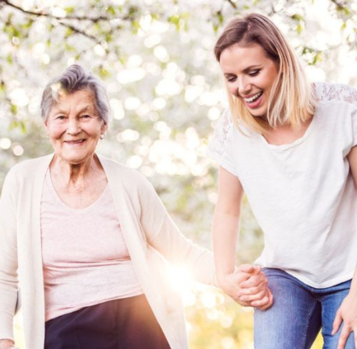 personal care (5) A young caregiver walks outdoors beside a senior woman on a sunny day, with trees surrounding them. They hold hands and laugh together, sharing a joyful moment that reflects compassionate personal care in Gulf Shores.