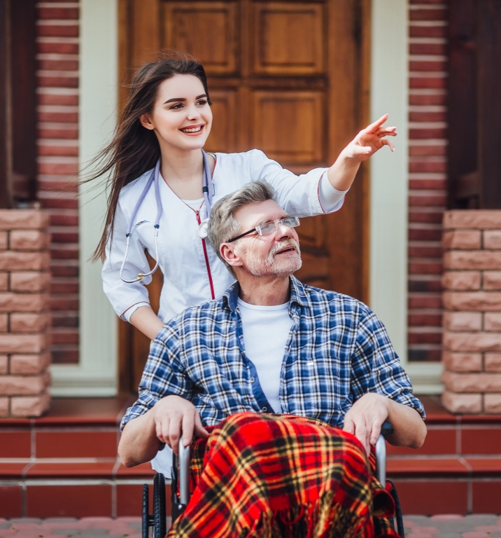 A woman caregiver with dark hair, pushes a veteran in wheel chair out side as she points to something in the distants.