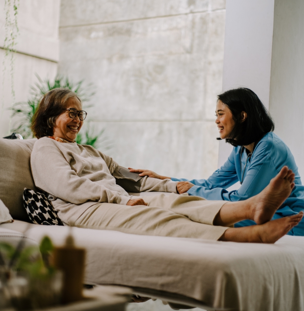 A smiling nurse in blue scrubs sits beside an elderly woman who is comfortably lying on a couch, engaging in warm conversation and holding her hand. This image reflects compassionate and personalized Skilled Nursing Care in Gulf Shores.