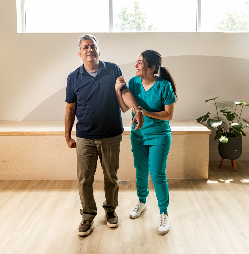 A nurse in teal scrubs assists a smiling middle-aged man as he walks indoors, offering physical support and encouragement. This image represents compassionate and personalized Skilled Nursing Care in Gulf Shores.
