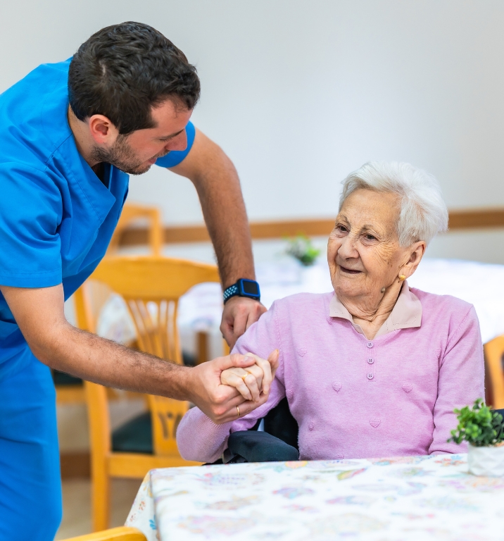 A caring nurse in blue scrubs offering Add-on Nursing Support Services while assisting a smiling elderly woman at a table, showing trust and compassion.