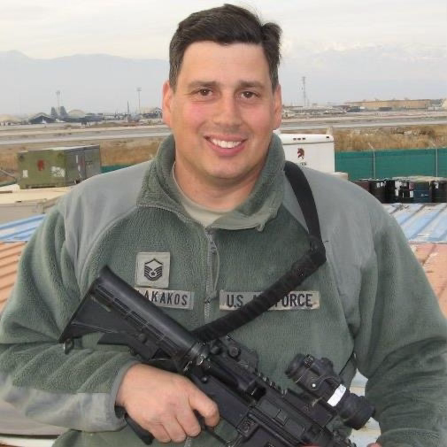 Honoring Master Sergeant Pete J. Nikolakakos, U.S. Air Force Veteran A smiling U.S. Air Force Master Sergeant Pete J. Nikolakakos in uniform holding a service rifle, standing outdoors near a base with mountains in the background.