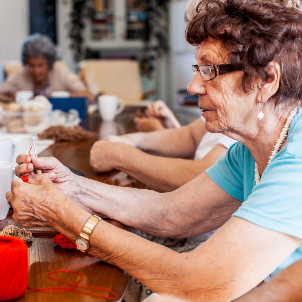 A group of senior women sitting together at a table, knitting and engaging in a creative activity in a warm, social environment.
