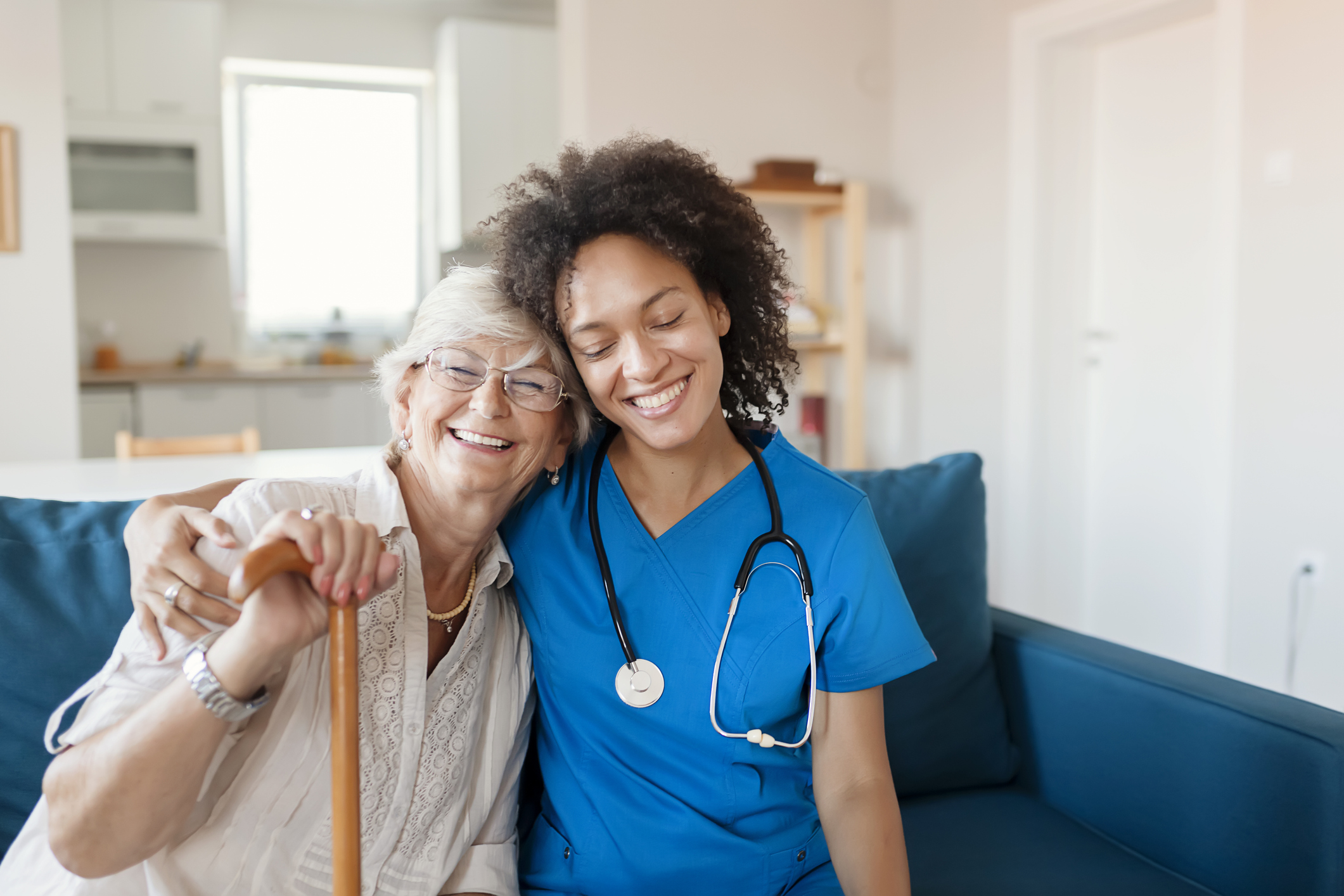 A nurse in blue scrubs provides care to an elderly man lying in bed, gently checking his chest with a stethoscope. The scene takes place in a bright, home-like setting, illustrating compassionate Skilled Nursing Care in Gulf Shores.