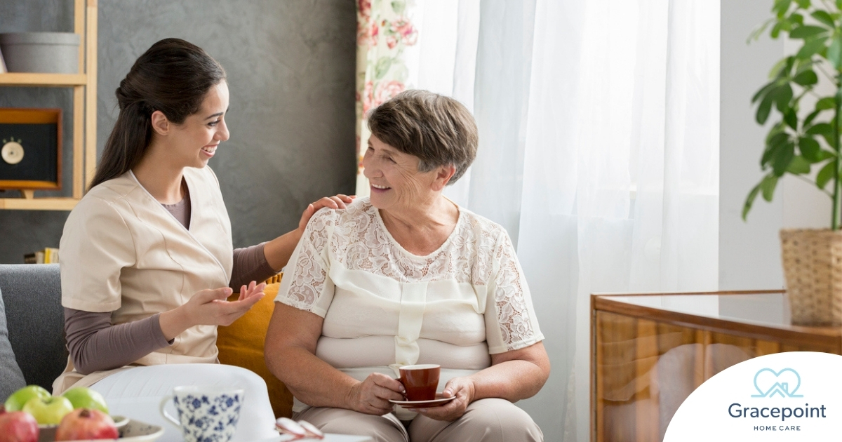Smiling caregiver with her hand on an older woman’s shoulder as they share tea on the couch, showing home care vs assisted living